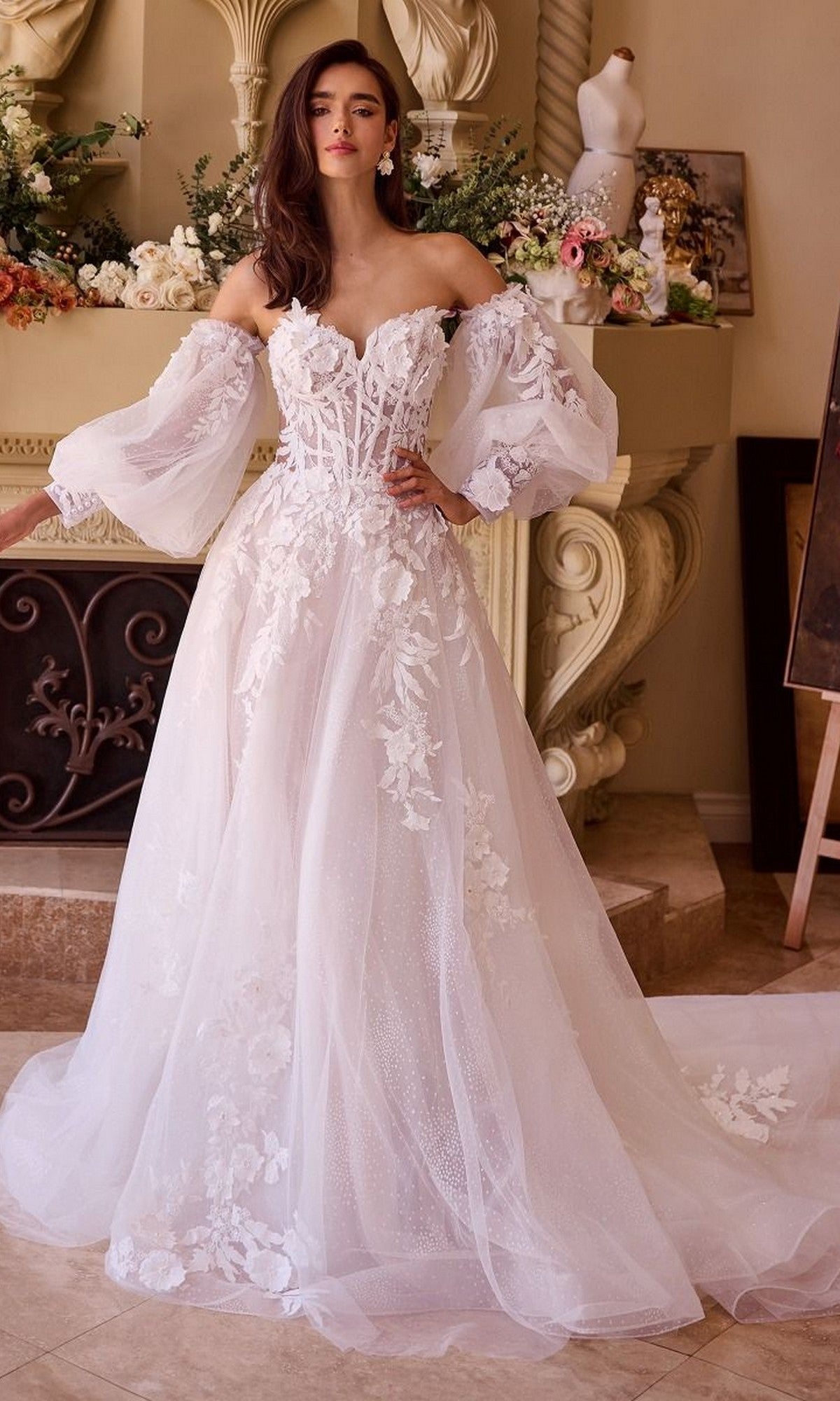 A woman wearing the Andrea and Leo WL018 long wedding dress with a strapless lace design, sweetheart neckline, and sheer puff sleeves stands indoors by a floral-decorated fireplace.